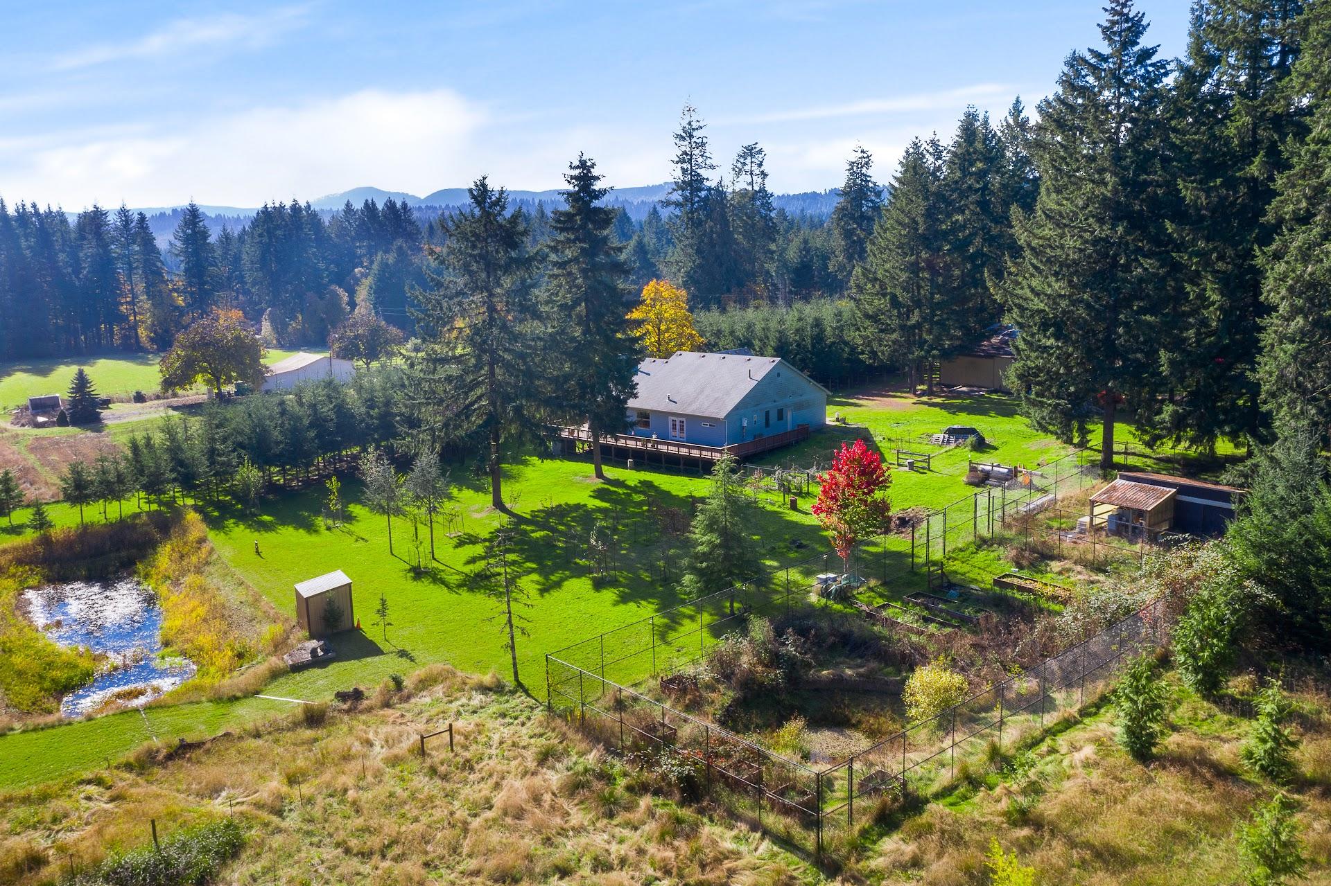 Aerial view of a rural Oregon property featuring a single-story home on acreage, fenced garden areas, a small pond, and surrounding evergreen trees with mountain views in the distance.