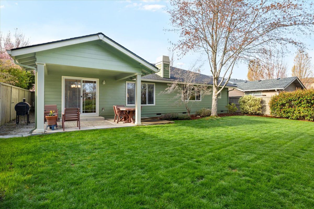 Backyard of a single-story green home with a covered patio, large lawn, fence, trees, and surrounding landscaping.