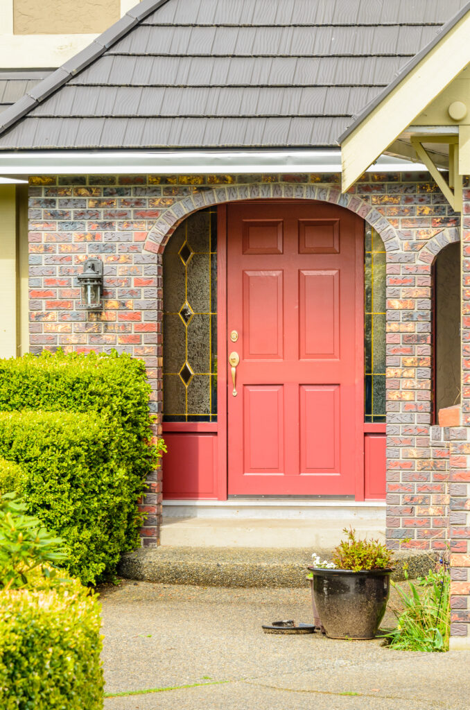 Front entry of a home with a red door, brick archway, and decorative glass side panels, with shrubs and a potted plant along the walkway leading up to the entrance.